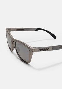 Ochelari de soare Oakley cu un cadru metalic gri, lentile întunecate, design geometric și vârfuri texturate pentru aderență și confort.