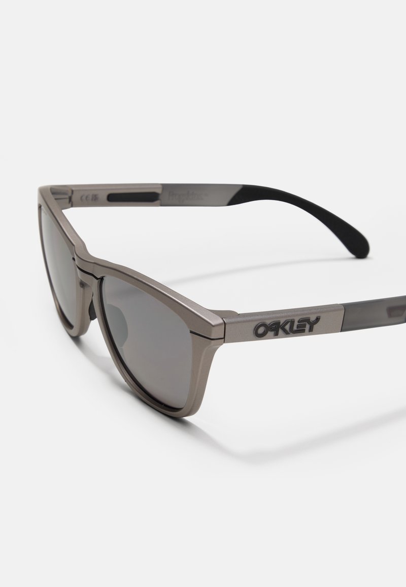 Gafas de sol Oakley con un marco metálico gris, lentes oscuros, diseño geométrico y puntas de las orejas texturizadas para un mejor agarre y comodidad.