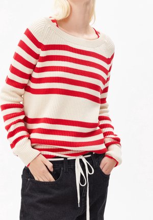 Henkilö, joka pukeutunut punavalkoraidalliseen neulepaitaan ja jolla on kädet mustien farkkujen taskuissa, näyttää keskivartalon ja kaulan valkoista taustaa vasten.