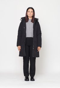 Cappotto lungo nero con cappuccio, con chiusura lampo frontale, tasche laterali e un maglione grigio testurizzato sotto. Indossato con pantaloni e scarpe neri.