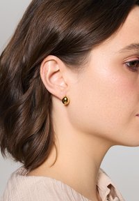 Złota, okrągła kolczyk o gładkiej, refleksyjnej powierzchni, umieszczona na uchu modelki. Modelka ma średniej długości ciemne włosy i jasną bluzkę.