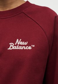 Μακρυμάνικη μπλούζα φούτερ σε κεραμιδί χρώμα με ριμπ γιακά, εμφανίζει λευκό κείμενο λογότυπου "New Balance" στο στήθος, δείχνοντας το άνω μέρος του κορμού και τη ραφή του ώμου.