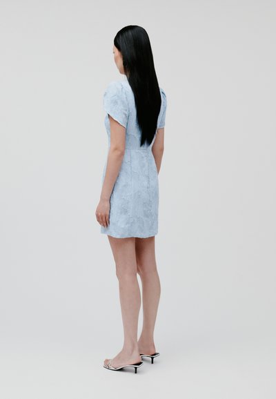 Hellblaues Kleid mit strukturiertem Blumenmuster, kurzen Puffärmeln, tailliertem Oberteil und einer Länge, die über dem Knie endet. Trägt dazu klare High Heels.