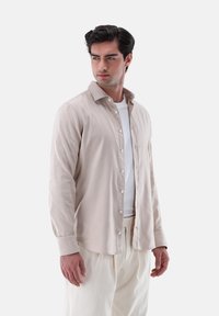 Camicia a maniche lunghe color beige chiaro con chiusura a bottoni, texture morbida, colletto classico e polsini singoli, indossata sopra una t-shirt bianca.