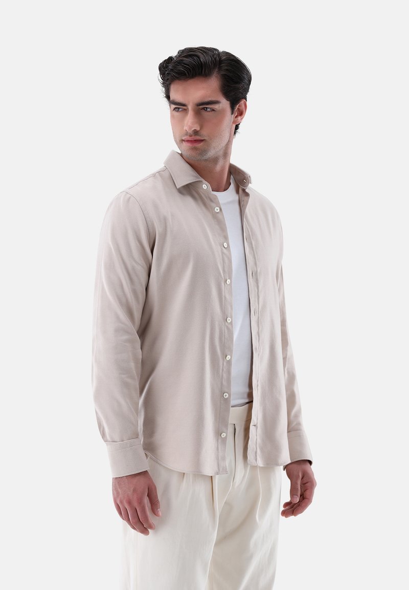Camicia a maniche lunghe color beige chiaro con chiusura a bottoni, texture morbida, colletto classico e polsini singoli, indossata sopra una t-shirt bianca.