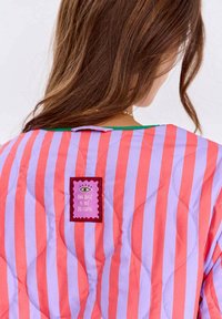 Kobieta z długimi brązowymi włosami w pikowanej kurtce w pionowe różowe i fioletowe paski oraz z naszywką z napisem "Najlepsze jeszcze przed nami."