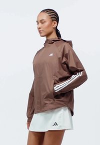 Braune Kapuzenjacke mit weißen drei Streifen, die einen lockeren Schnitt und seitliche Taschen aufweist, kombiniert mit einem weißen Rock. Glatte Textur.