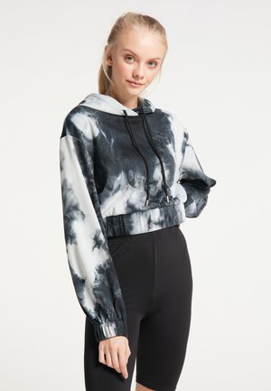 Młoda kobieta o blond włosach, ubrana w czarno-białą krótka bluzę z kapturem typu tie-dye oraz czarne spodenki rowerowe, stoi na jednolitym tle.