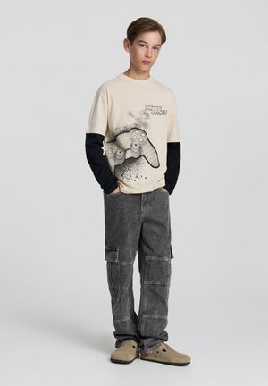 T-shirt di cotone beige con grafica di un controller di videogioco, sovrapposta a una maglietta nera a maniche lunghe. Pantaloni cargo grigi e scarpe beige senza lacci.
