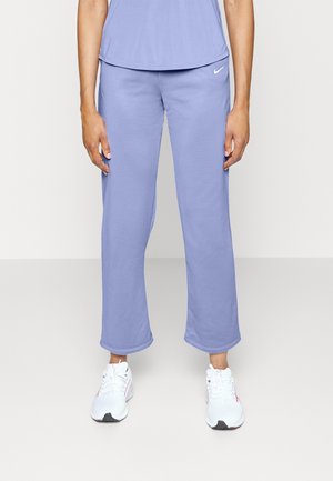 Persona vistiendo pantalones deportivos Nike de color púrpura claro y una parte superior a juego, de pie con los brazos relajados, y zapatos deportivos blancos sobre un fondo simple.