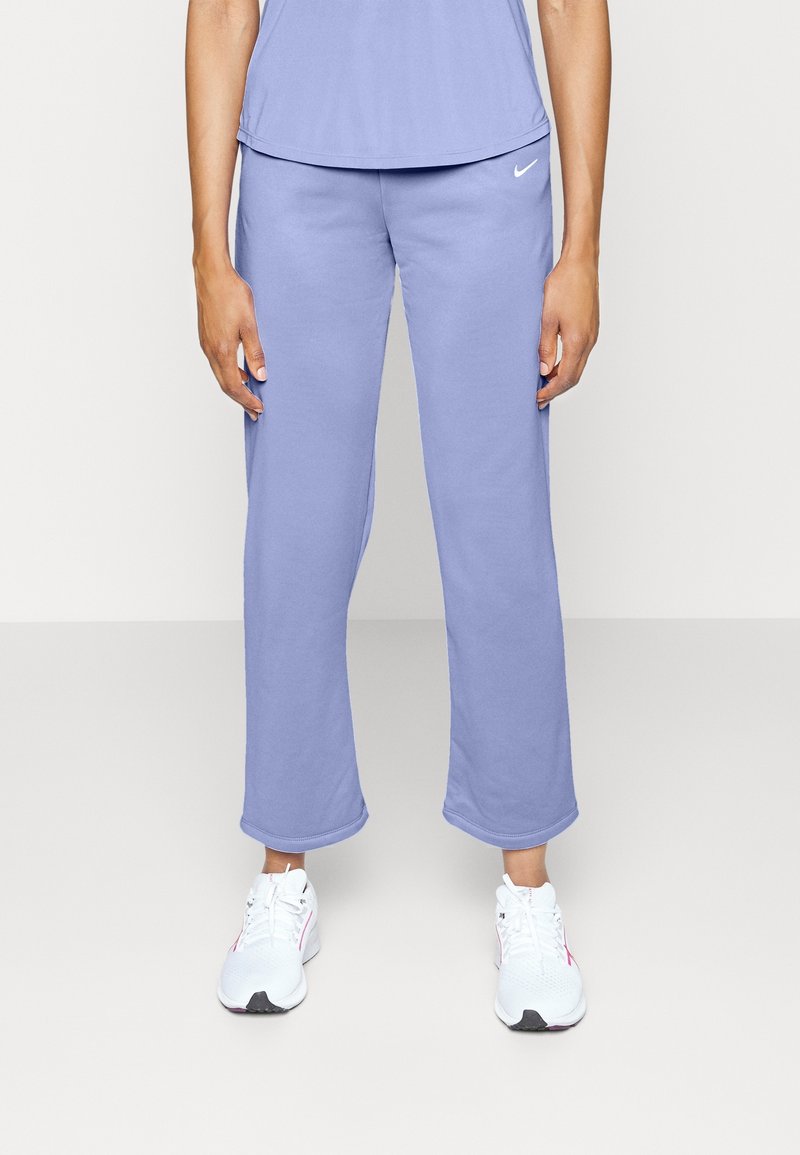 Person som bär ljuslila Nike-joggingbyxor och matchande överdel, står med avslappnade armar och vita sportskor mot en neutral bakgrund.