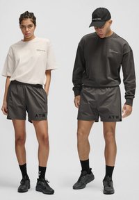 Två modeller bär matchande avslappnade kläder: en ljus beige stor t-shirt och en mörk sweatshirt med svarta shorts, som har "ATW"-logotypen i svart.