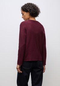 Burgunderfarbener Pullover mit relaxed Fit, gerippter Textur und langen Ärmeln. Der Saum endet knapp über dem Bund einer dunklen Jeans.