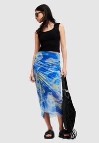 Διάφανη φούστα με μπλε και κίτρινο σχέδιο και draping, συνδυασμένη με μαύρο αμάνικο τοπ και μαύρες σανδάλες slide, κρατώντας μια μαύρη πλεγμένη τσάντα.