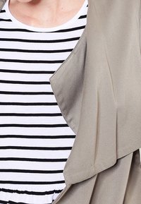 Chaqueta envolvente gris claro sobre una camisa a rayas blanca y negra. La chaqueta tiene un tejido suave, un diseño fluido y un frente abierto. Las rayas son horizontales.