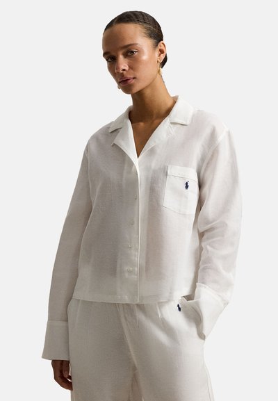 Vrouw met gevlochten haar draagt een wit overhemd met knopen en bijpassende broek met Polo Ralph Lauren-logo op de zak en het dijbeen.