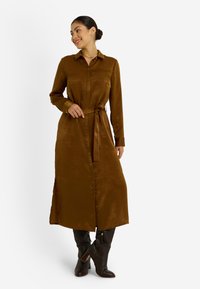 Vestido marrón de manga larga hecho de un tejido suave y texturizado, con cuello, botones y un cinturón atado a la cintura. Combinado con botas hasta la rodilla.