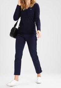 Kobieta ubrana w granatową bluzkę z długim rękawem i dekoltem w serek, dopasowane spodnie, białe trampki, trzymająca czarną torbę na ramię, stojąca na jednolitym tle.