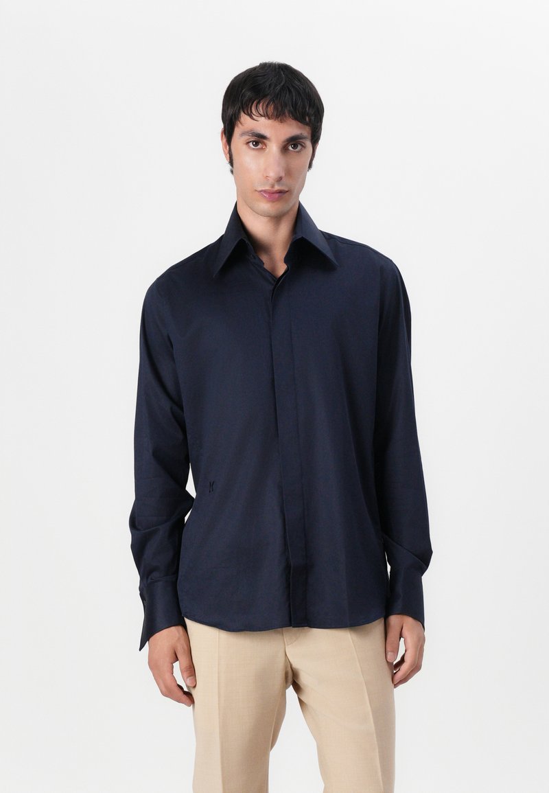 Cămașă polo bleumarin, din țesătură netedă, cu guler clasic și mâneci lungi, având un detaliu mic cu logo aproape de tiv.