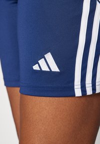 Marine atletiske shorts med hvite tre-striper og Adidas-logo. Laget av glatt, elastisk stoff. Nærbilde av benområdet.