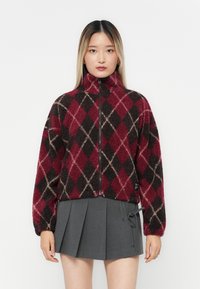 Rød og svart fleecejakke med argyle-mønster, høy krave og glidelås foran. Modellen har på seg et grått plissé skjørt som fremhever lagdelte teksturer.