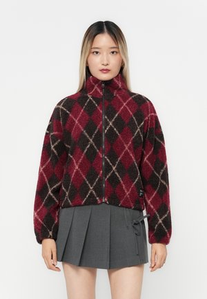 Red and black fleece jacket with an argyle pattern, high collar, and zip front. The model wears a gray pleated skirt, showcasing layered textures.