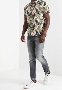 Camicia a maniche corte con bottoni, caratterizzata da un vivace motivo di foglie in verde, giallo e rosso. Indossata con jeans grigi sbiaditi e sneakers bianche.