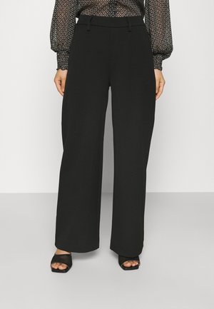 Frau trägt schwarze, hoch taillierte, weit geschnittene Hose, halbtransparente, gemusterte Bluse und schwarze Sandalen mit offenen Zehen, steht vor einfachem Hintergrund.