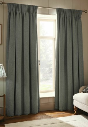 La luce filtra dolcemente attraverso una finestra con cornice bianca e tende chiuse verde-grigio in un soggiorno accogliente con pavimento in legno e divano beige.