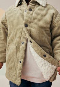 Veste en velours côtelé vert olive avec col doublé en sherpa, dotée de boutons-pression noirs et de poches latérales, partiellement déboutonnée pour montrer l'intérieur.