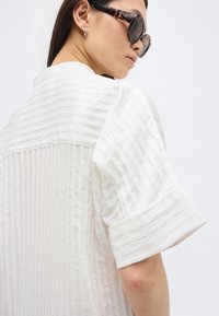 Γυναίκα με γυαλιά ηλίου τύπου χελώνας και υφασμάτινο λευκό τοπ με φαρδιά μανίκια, στραμμένη στο πλάι μπροστά σε απλό λευκό φόντο.