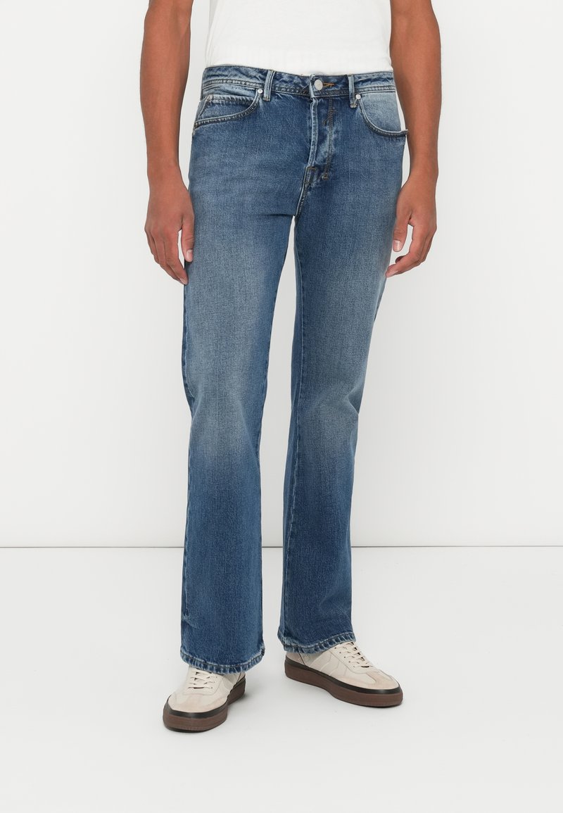 Φθαρμένα τζιν σε μπλε απόχρωση με ίσια γραμμή, σχέδιο πέντε τσεπών και κουμπωτή μπροστινή τσέπη. Φορεμένα με κρέμ σα Sneakers, σε λευκό φόντο.