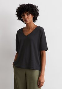 Black cotton V-neck t-shirt with short sleeves, featuring a relaxed fit and a smooth texture. Paired with green wide-legged trousers.