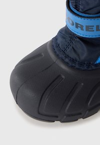 Bota negra impermeable con suela de goma texturizada, parte superior de nailon azul y correa azul ajustable. Presenta detalles estriados para mayor agarre.