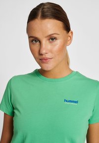 Grøn bomulds t-shirt med korte ærmer, rund halsudskæring, der har et blåt logo "hummel SPORTSWEAR" på venstre brystområde.