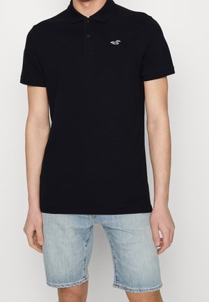 Schwarzes Baumwoll-Polohemd mit Kragen, drei Knöpfen und kleinem Logo auf der Brust. Getragen mit hellblauen Jeansshorts.