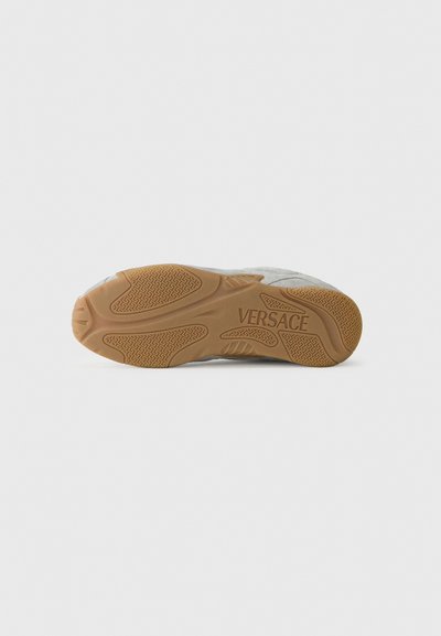 Versace sneakersula, tillverkad av ett gummimaterial i ljusbrunt med ett strukturerat mönster, och varumärket präglat. Slät grå ovandel synlig.