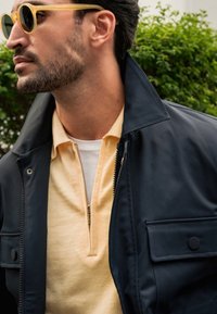 Chaqueta de marina con un alto cuello y un cierre frontal con cremallera, combinada con un polo amarillo claro con media cremallera, todo ello en un entorno de vegetación.