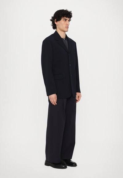 Sacou de marină cu un design structurat, prevăzut cu un singur nasture, asociat cu pantaloni largi, cu picioare ample și pantofi negri lustruiți.