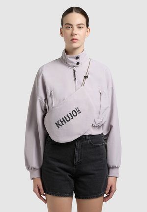 Woman wearing a pale purple zip-up jacket and black shorts with a large KHUJO 2001 crossbody bag.