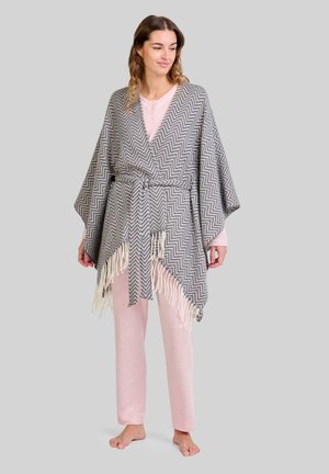Poncho con patrón de chevron en gris y blanco, con bordes con flecos, ajustado a la cintura con un lazo. Lleva puesto un pantalón rosa.