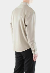 Mann iført lys beige skjorte med lange ermer og svarte bukser, vist fra ryggen mot en helt hvit bakgrunn.