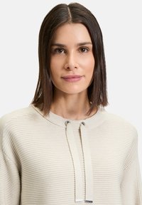 Beige gestrickter Pullover mit einem strukturierten, gerippten Design. Verfügt über einen weiten Ausschnitt mit Kordelzügen und silbernen Hardware-Akzenten.