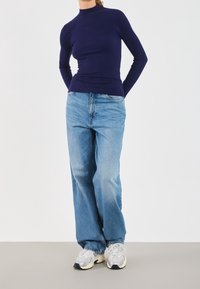 Person som bär en marinblå ribbad långärmad tröja, ljusblå vida jeans och vita sneakers, står med händerna bakom ryggen.