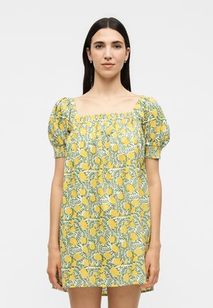 Donna con lunghi capelli scuri indossa un vestito corto giallo floreale con maniche a sbuffo e scollo quadrato, in piedi davanti a uno sfondo semplice.