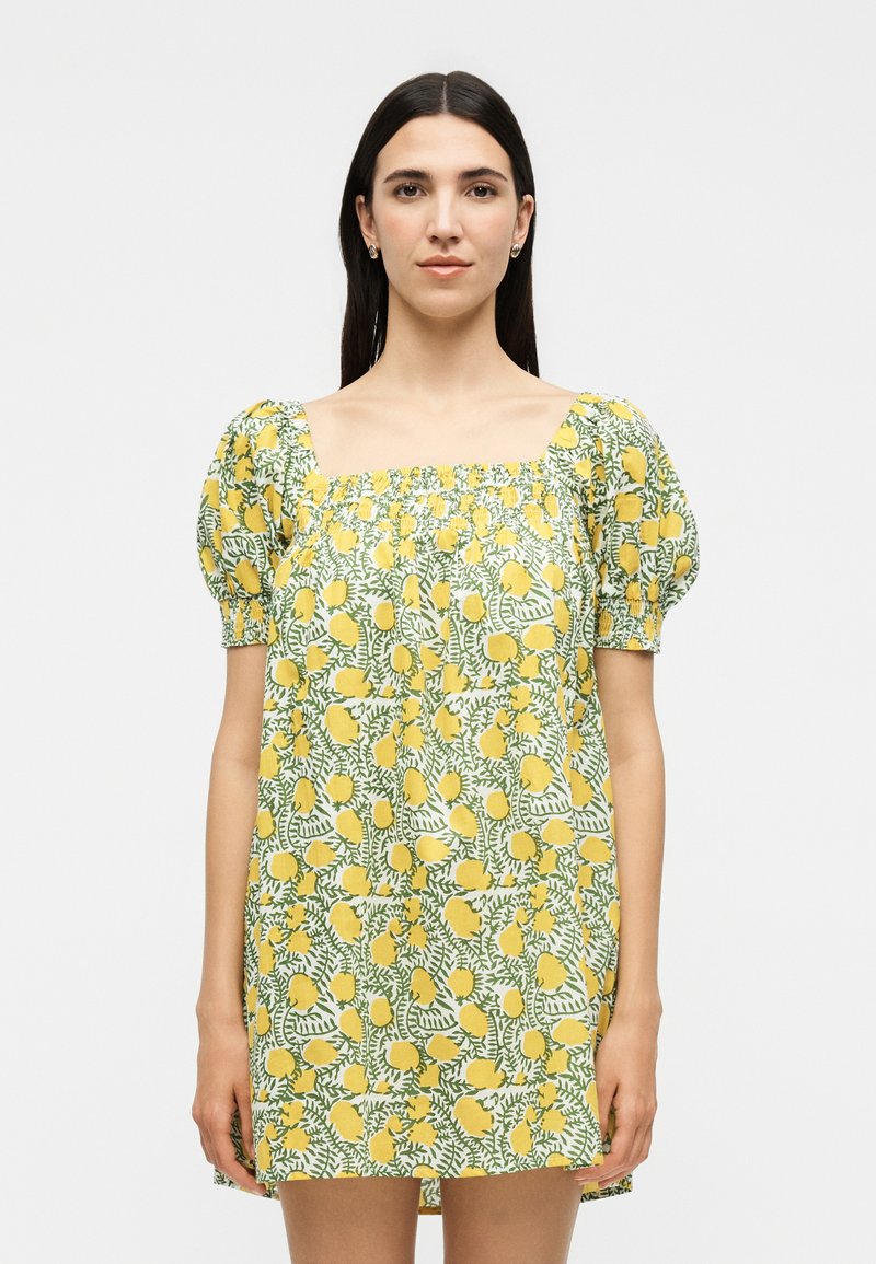 Femme aux longs cheveux foncés porte une courte robe jaune fleurie avec des manches bouffantes et un décolleté carré, debout devant un fond uni.