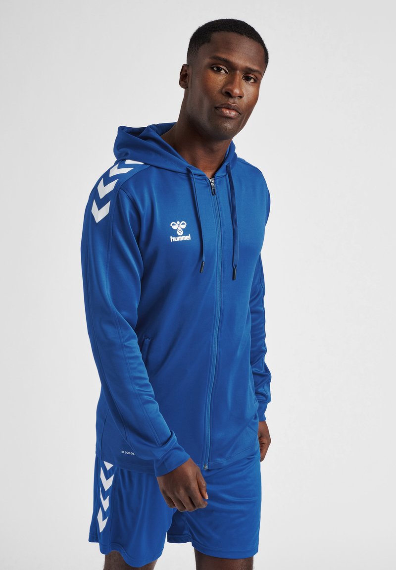 Felpa blu con zip e pantaloni corti abbinati realizzati in tessuto morbido. La felpa presenta motivi a chevron bianchi sulle maniche e un logo sul petto.