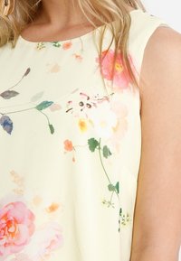 Květinová halenka z jemné žluté tkaniny. Má růžové a zelené květinové vzory, kulatý výstřih a bezrukávový střih s volným střihem.