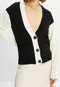 Czarnobiały sweter na drutach z głębokim dekoltem w serek, o prążkowanej fakturze, z dwoma przednimi kieszeniami i dużymi czarnymi guzikami. Rękawy są długie i kontrastowe.