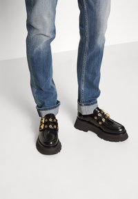 Zapatos de cuero negro con detalles en hardware dorado, suelas gruesas y dos diseños de correas. Llevados con jeans azules arremangados.